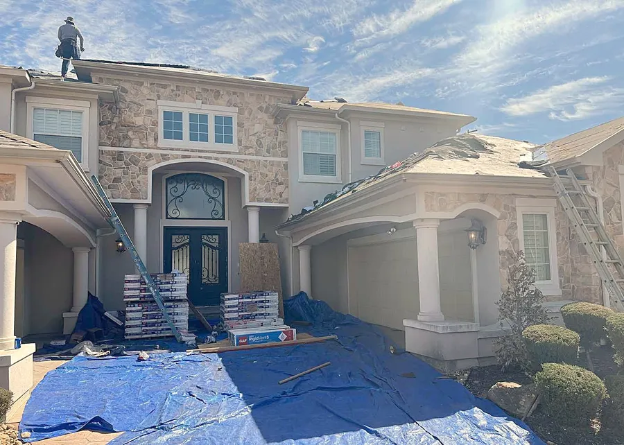 A re-roof under way on a stone and stucco home, tarps down and GAF materials stacked by the door